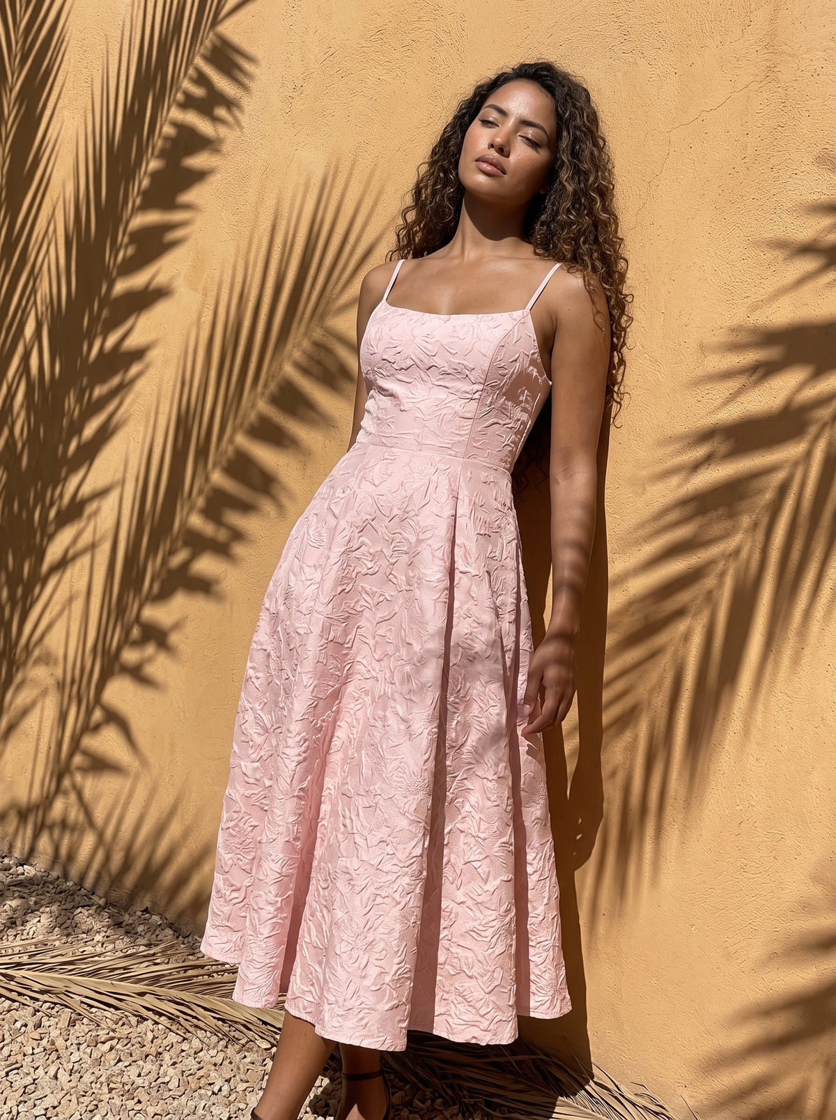 Model in a blush pink jacquard midi dress against a sunlit ochre wall